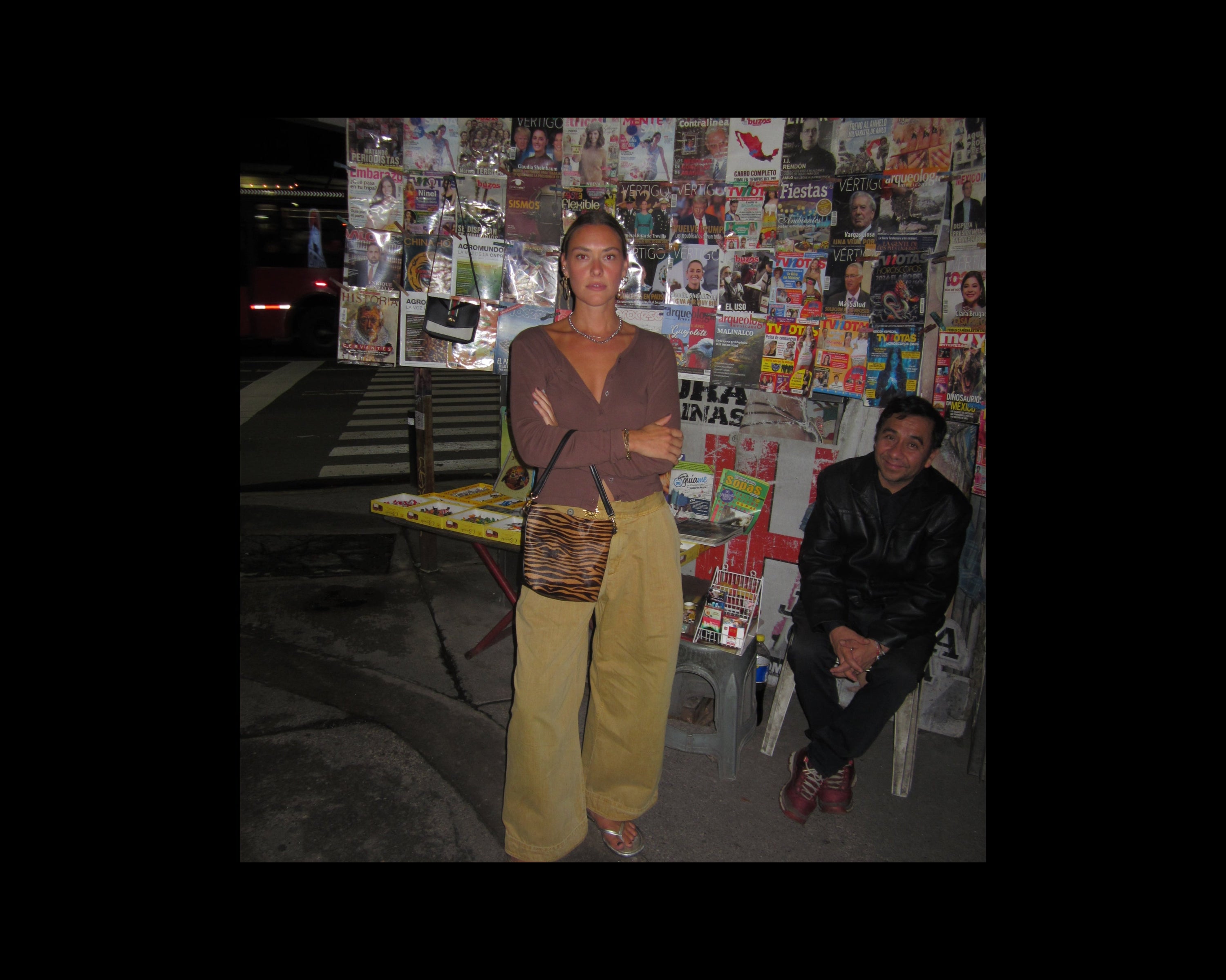 A woman stands in front of a street newspaper stand at night, wearing a brown wrap top, wide-leg beige pants, and carrying a brown and black zebra-patterned handbag. A man sits on a stool to the right against a wall covered with magazines and newspapers.